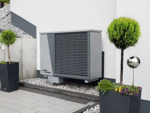 A new outdoor heat pump unit set on gravel beside a white wall with potted plants
