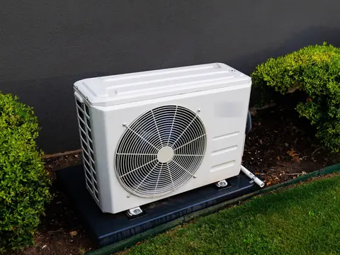 A compact mini split outdoor unit on a pad by a hedge