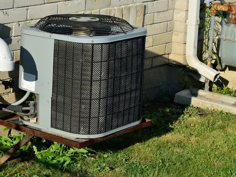 An older outdoor AC unit, weathered, on a rusted stand by the house